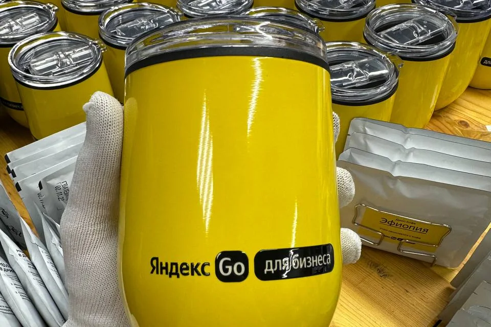 Жёлтая термокружка «Яндекс Go для бизнеса» в руке, за ней — тираж таких же