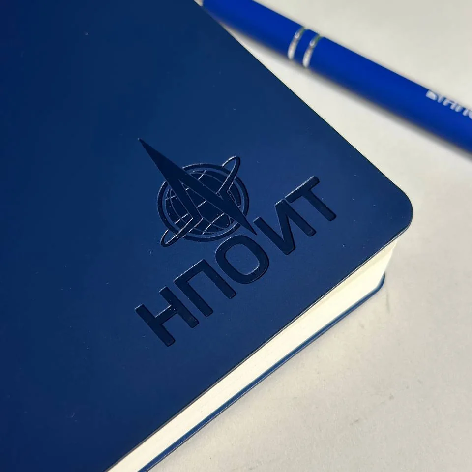 Синий блокнот с тиснёным логотипом НПО ИТ, рядом ручка